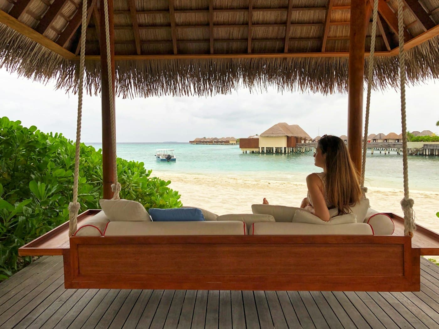 Woman relaxing on a swing bed in a thatched beach cabana, overwater villas beyond